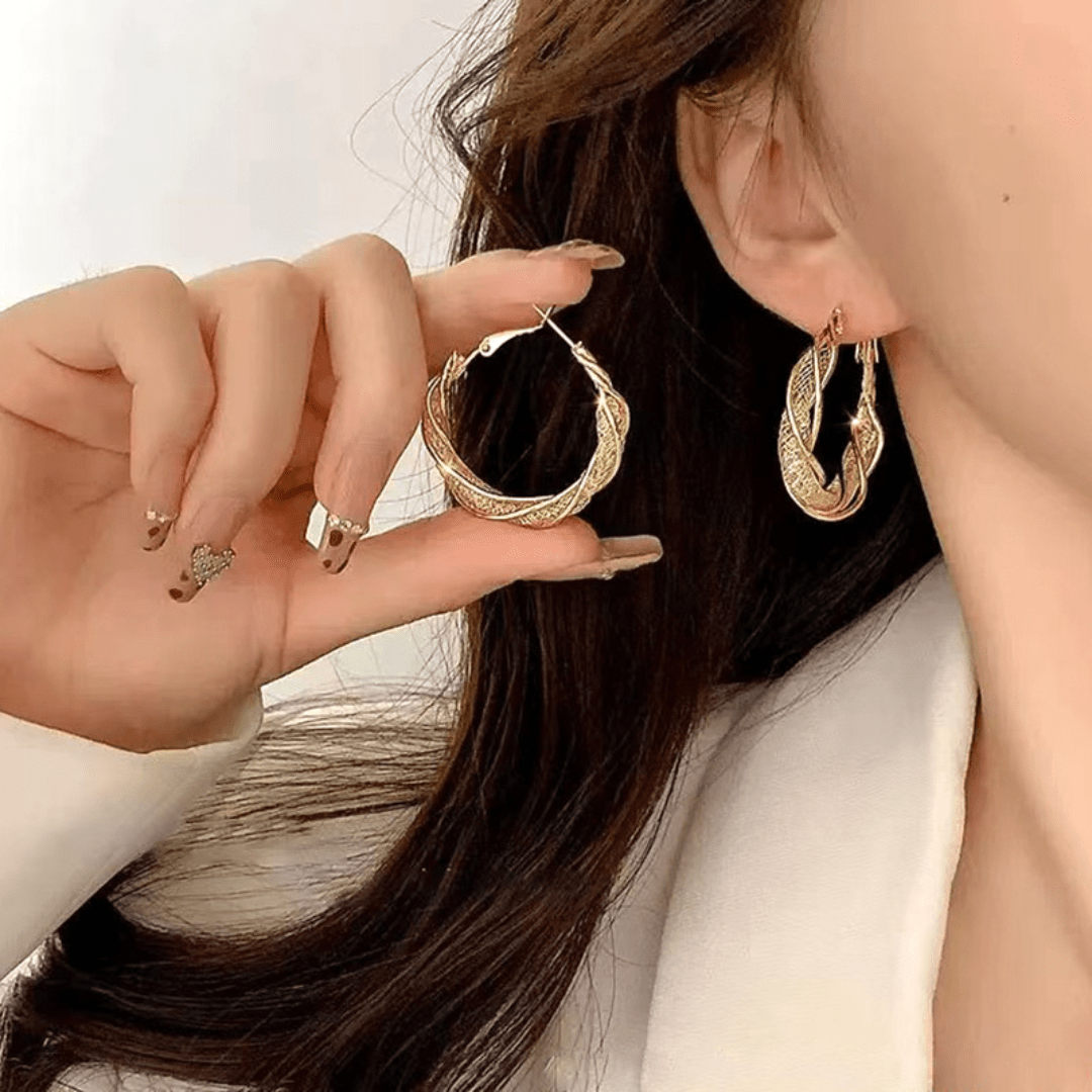 Una mujer de pelo castaño largo y uñas desnudas cuidadas lleva una americana blanca y aros dorados, y sostiene los Aretes colgantes en espiral cerca de la cara. Sus uñas lucen un delicado nail art.