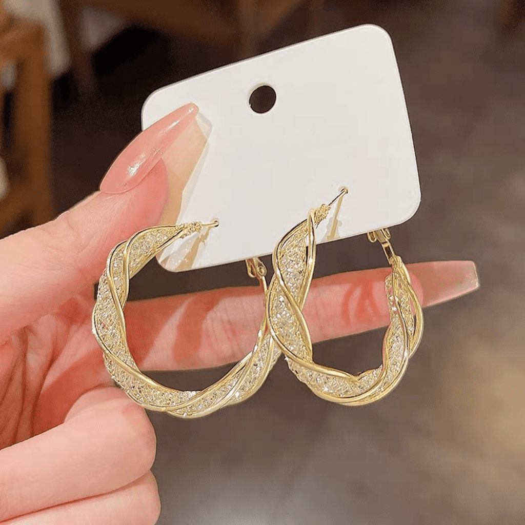 Una mano con las uñas largas y cuidadas sostiene una tarjeta blanca que muestra Aretes colgantes en espiral, elaborados en acero inoxidable dorado con piedras brillantes incrustadas.