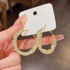 Una mano con las uñas largas y cuidadas sostiene una tarjeta blanca que muestra Aretes colgantes en espiral, elaborados en acero inoxidable dorado con piedras brillantes incrustadas.