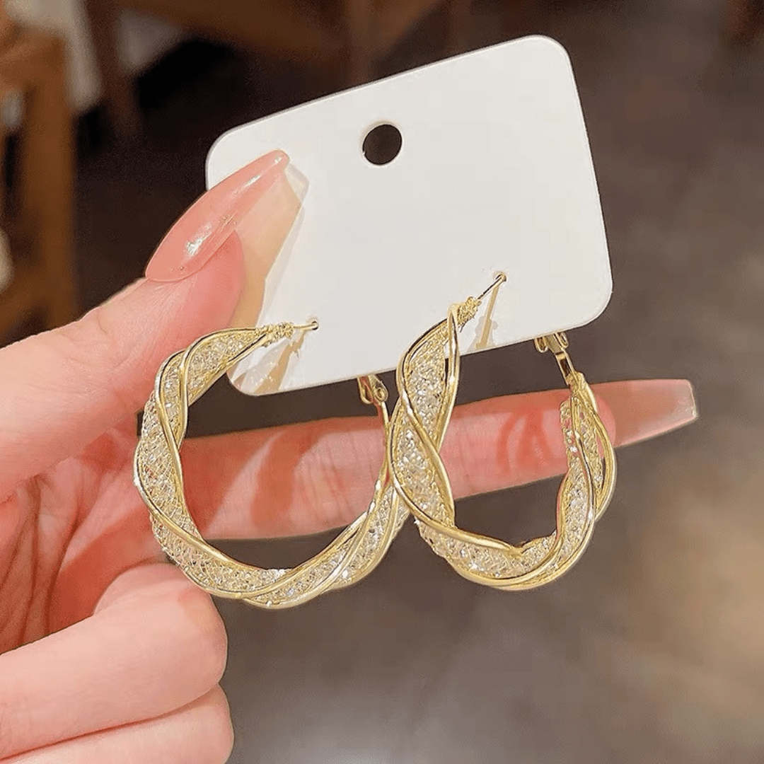 Una mano con las uñas largas y cuidadas sostiene una tarjeta blanca que muestra Aretes colgantes en espiral, elaborados en acero inoxidable dorado con piedras brillantes incrustadas.