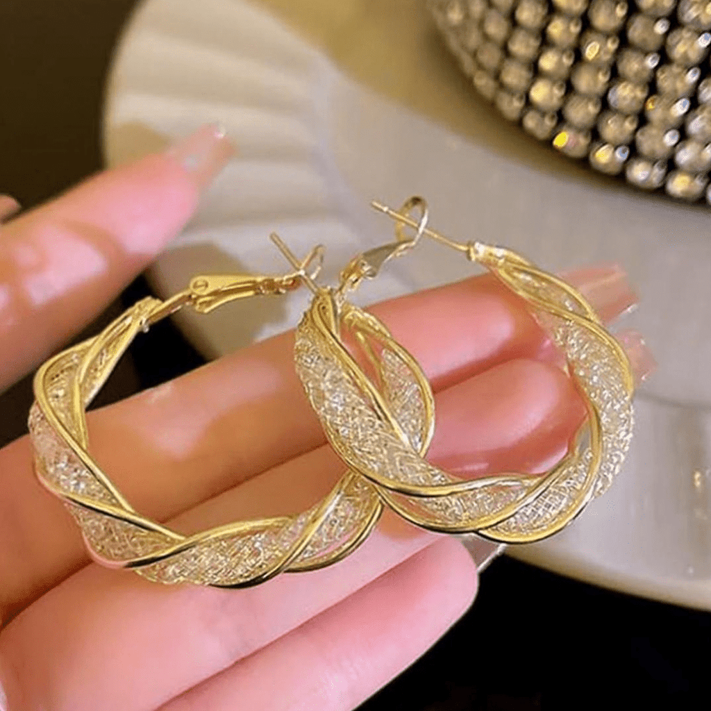 Una mano muestra Aretes colgantes en espiral, que lucen hebras de plata retorcida acentuadas con suaves bandas doradas. Al fondo, un objeto decorativo de abalorios aparece suavemente difuminado.