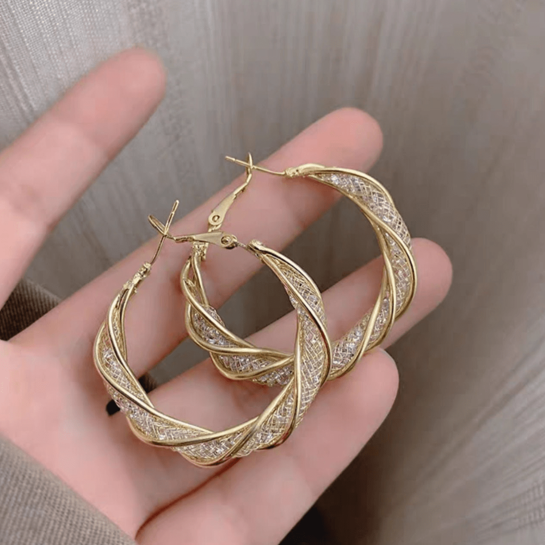 Una mano sujeta Aretes colgantes en espiral, con aros de oro retorcidos con un brillante diseño de celosía en tonos dorados y plateados mezclados, elaborados en acero inoxidable dorado para un aspecto texturizado y elegante.