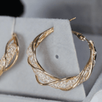 Un primer plano de Aretes colgantes en espiral, con intrincados aros de oro retorcidos con piedras brillantes, elaborados en acero inoxidable dorado y expuestos en un soporte de joyería gris claro.
