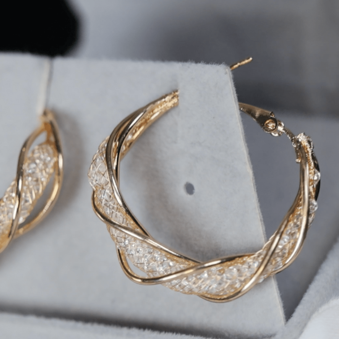 Un primer plano de Aretes colgantes en espiral, con intrincados aros de oro retorcidos con piedras brillantes, elaborados en acero inoxidable dorado y expuestos en un soporte de joyería gris claro.