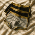 Tres pares de Boxers icónicos -dos negros y uno verde oliva, con cintura y ribetes elásticos dorados- se exhiben sobre tela beige bajo una suave luz natural.