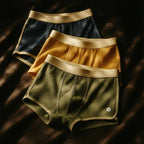 Tres pares de Boxers icónicos para hombre en tejido acanalado -azul oscuro, amarillo mostaza y verde oliva- con cinturilla beige, ordenados sobre una superficie oscura bajo una luz tenue.