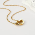 A Caja Corazón con rosa eterna y un collar corazón personalizado, con dos colgantes en forma de corazón entrelazados, uno grabado con el nombre Merel, expuesto sobre una superficie blanca.