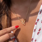 Una persona con las uñas rojas señala un collar dorado en forma de corazón, con un top floral blanco: un regalo ideal con la Caja Corazón con rosa eterna y collar corazón personalizado.