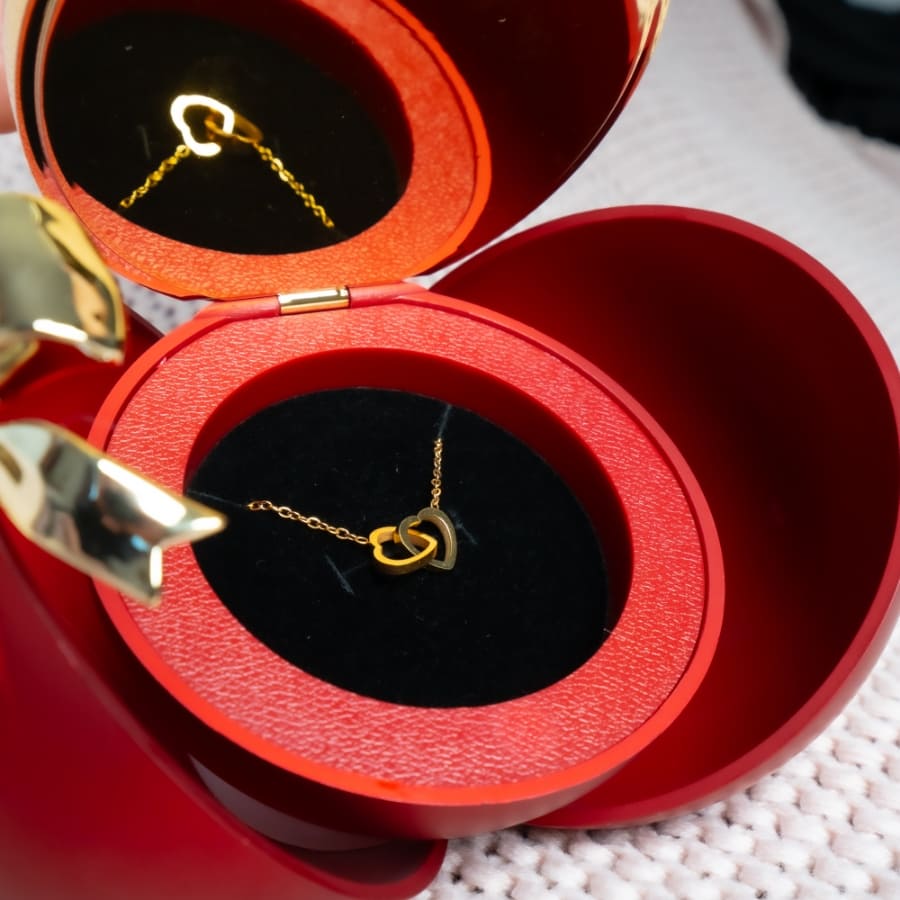 Un collar de corazón personalizado con un colgante de oro se exhibe en la Caja Corazón, que presenta una rosa eterna y descansa dentro de un joyero rojo con forma de manzana forrado con tela negra y roja para una presentación encantadora.