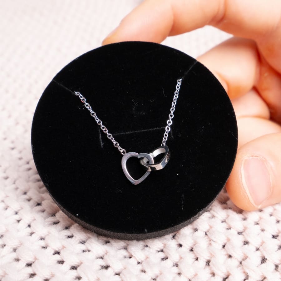 Una mano sostiene una Caja Corazón negra con rosa eterna y collar corazón personalizado, que muestra un collar de plata con dos colgantes de corazón entrelazados sobre una superficie de punto color crema.