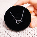 Una mano sostiene una Caja Corazón negra con rosa eterna y collar corazón personalizado, que muestra un collar de plata con dos colgantes de corazón entrelazados sobre una superficie de punto color crema.