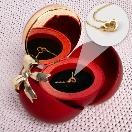 Una "Caja Corazón con rosa eterna y collar corazón personalizado" que incluye un collar dorado en forma de corazón con la inscripción "Meral" grabada, presentado en una caja de regalo roja con un lazo dorado, perfecta para regalar con un toque de rosa eterna.