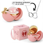 La Caja Corazón con rosa eterna y collar corazón personalizado incluye una caja rosa en forma de corazón con un lazo dorado, una rosa preservada, un portajoyas y un collar personalizado con dos anillos entrelazados para nombres grabados. La caja lleva la etiqueta "Love.
