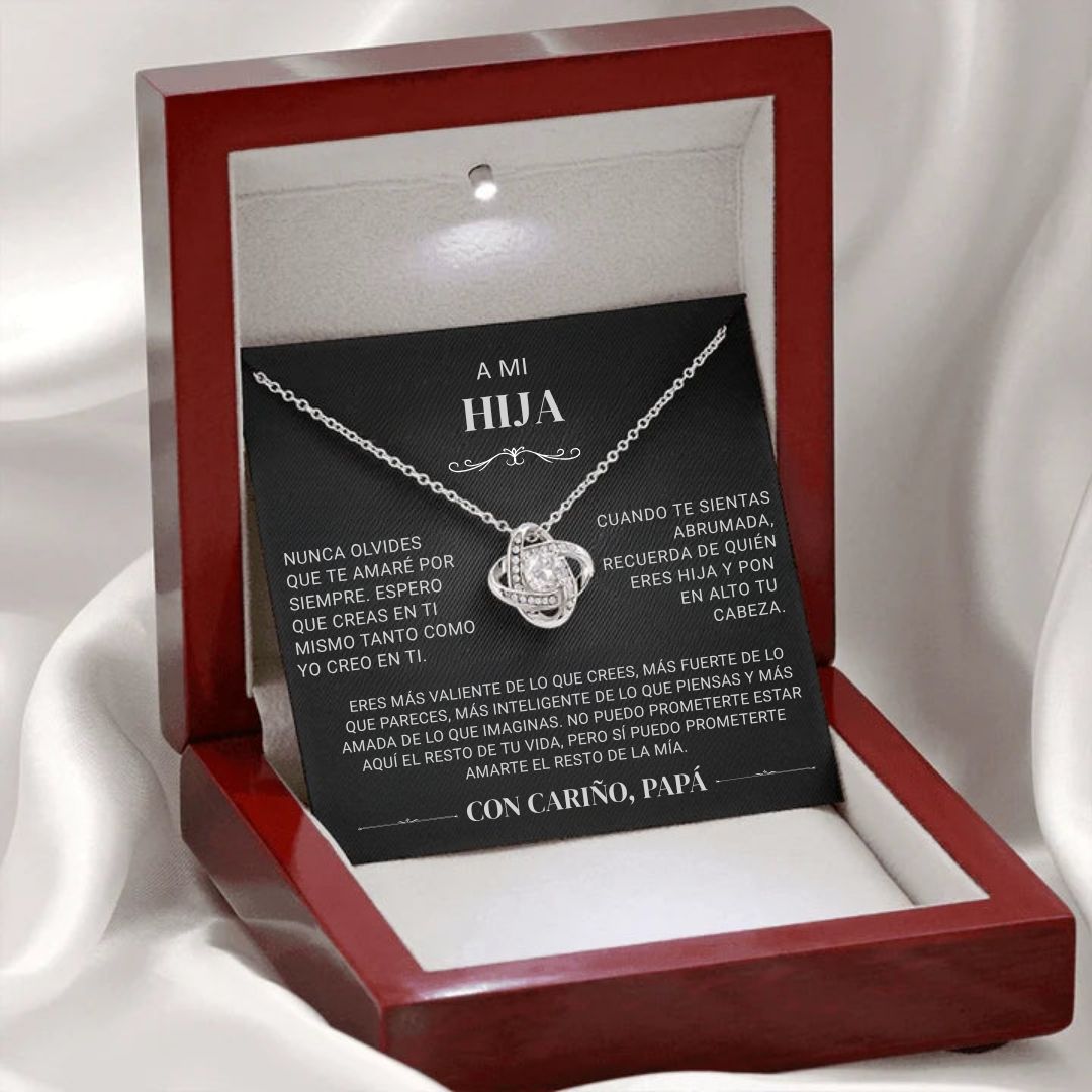 El collar "A mi hija" - Con cariño Papá, con un colgante de plata entrelazada, se presenta en una caja roja abierta con interior blanco. Detrás descansa una tarjeta en español con un sentido mensaje de un padre a su hija.