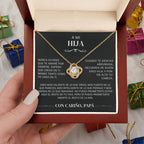 Una mano sostiene un joyero abierto que contiene el collar "A mi hija" - Con cariño Papá, junto con una tarjeta titulada "A mi hija" que contiene un sentido mensaje de un padre a su hija en español. Al fondo se ven coloridas cajas de regalo.