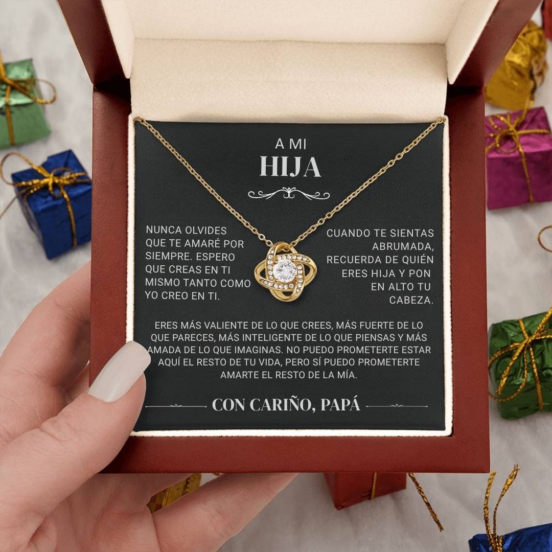 Una mano sostiene un joyero abierto que contiene el collar "A mi hija" - Con cariño Papá, junto con una tarjeta titulada "A mi hija" que contiene un sentido mensaje de un padre a su hija en español. Al fondo se ven coloridas cajas de regalo.