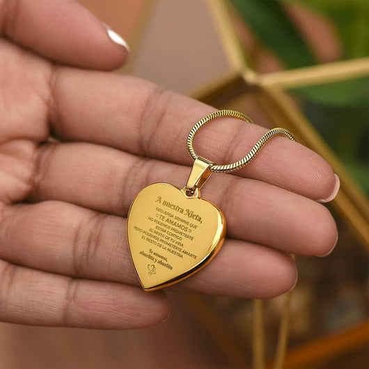 Una persona sostiene el Collar "A nuestra nieta" de corazón, un colgante dorado con mensaje grabado en español, perfecto como regalo para abuelos y nieta; cuelga de una cadena dorada y al fondo se ve una planta verde desenfocada.