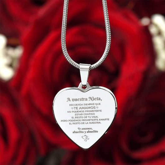 El "Collar 'A nuestra nieta' de corazón" es un collar personalizado de plata grabado en español, un regalo sentimental abuelos nieta con un sentido mensaje de los abuelos a su nieta. Las rosas rojas aparecen difuminadas en el fondo.