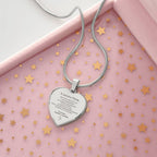 El collar "A nuestra nieta" de corazón, con mensaje grabado, reposa sobre una superficie rosa con confeti de estrellas doradas y está en una caja de joyería rosa-un regalo ideal para abuelos a su nieta.