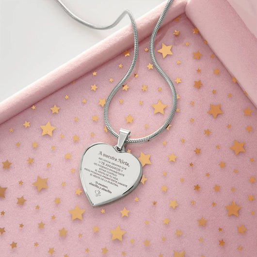 El collar "A nuestra nieta" de corazón, con mensaje grabado, reposa sobre una superficie rosa con confeti de estrellas doradas y está en una caja de joyería rosa-un regalo ideal para abuelos a su nieta.