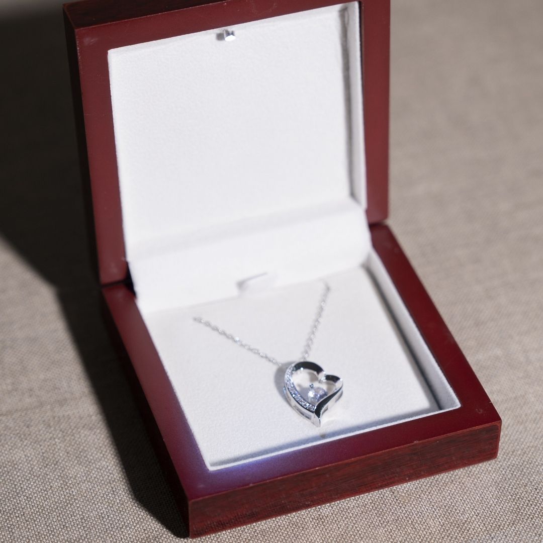 El collar Amor eterno - Para mi hija es un collar de acero inoxidable en forma de corazón con una piedra preciosa central, presentado en un joyero de madera abierto forrado con tela blanca sobre una superficie beige: un bonito regalo para su hija.