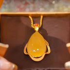 Un primer plano del Collar amuleto de piedra ágata amarilla: un colgante de ágata amarilla en forma de lágrima enmarcado en oro con una pequeña piedra roja encima, en una cadena de oro, presentado en un joyero marrón.
