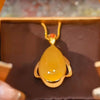 Un primer plano del Collar amuleto de piedra ágata amarilla: un colgante de ágata amarilla en forma de lágrima enmarcado en oro con una pequeña piedra roja encima, en una cadena de oro, presentado en un joyero marrón.