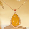 El Collar amuleto de piedra ágata amarilla es un diseño retro con un colgante ovalado de ágata amarilla enmarcado en oro texturizado y una pequeña piedra roja, irradiando energía positiva sobre fondo beige.