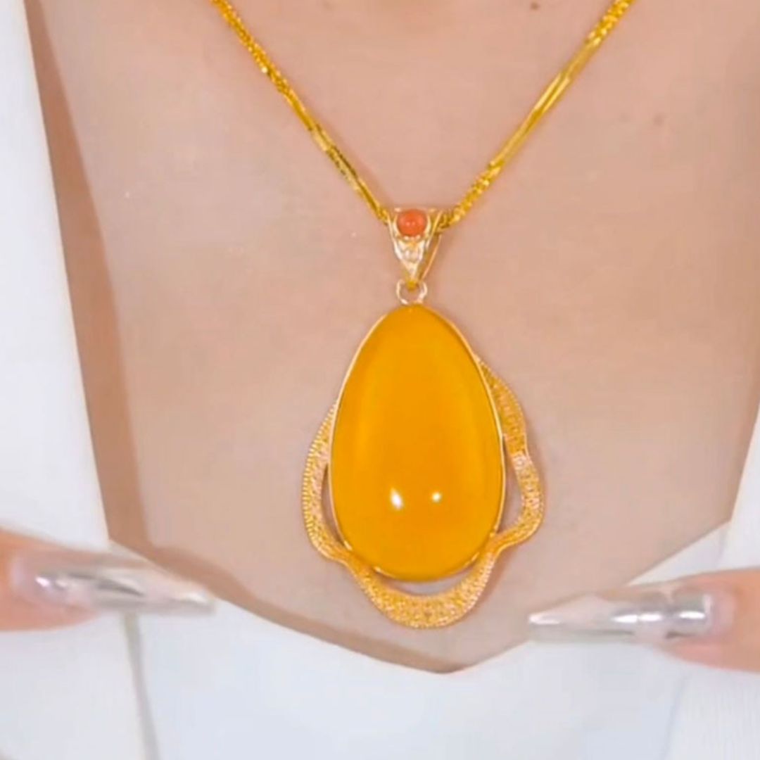 Un primer plano del Collar amuleto de piedra ágata amarilla, un collar retro de oro con un gran colgante ovalado liso de ágata amarilla y detalles dorados. Dos manos con largas uñas plateadas realzan su energía positiva sobre una prenda blanca.