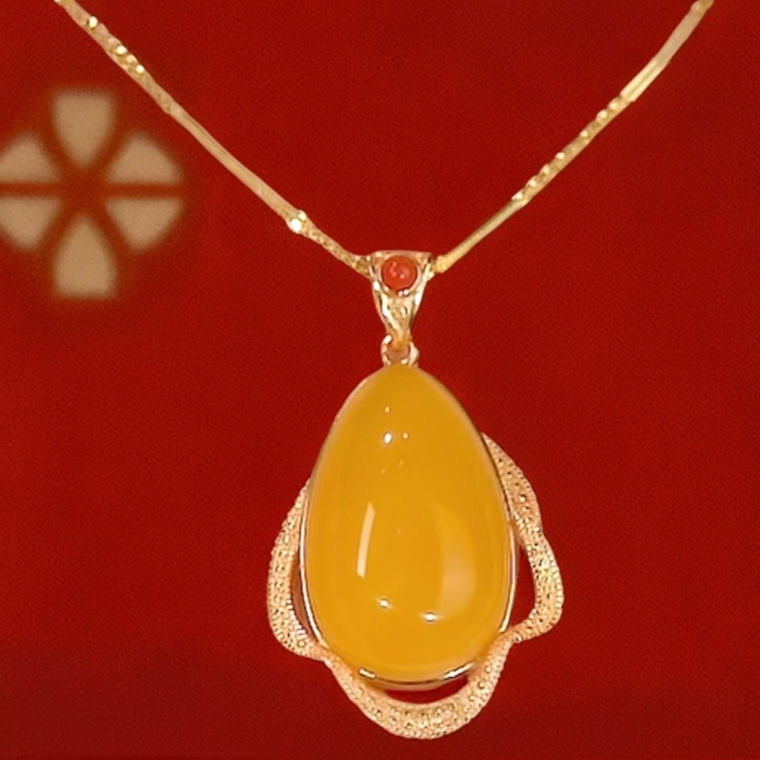 El Collar amuleto de piedra ágata amarilla, con dije en forma de gota rodeado por un marco dorado texturizado, irradia energía positiva sobre un fondo rojo vibrante.