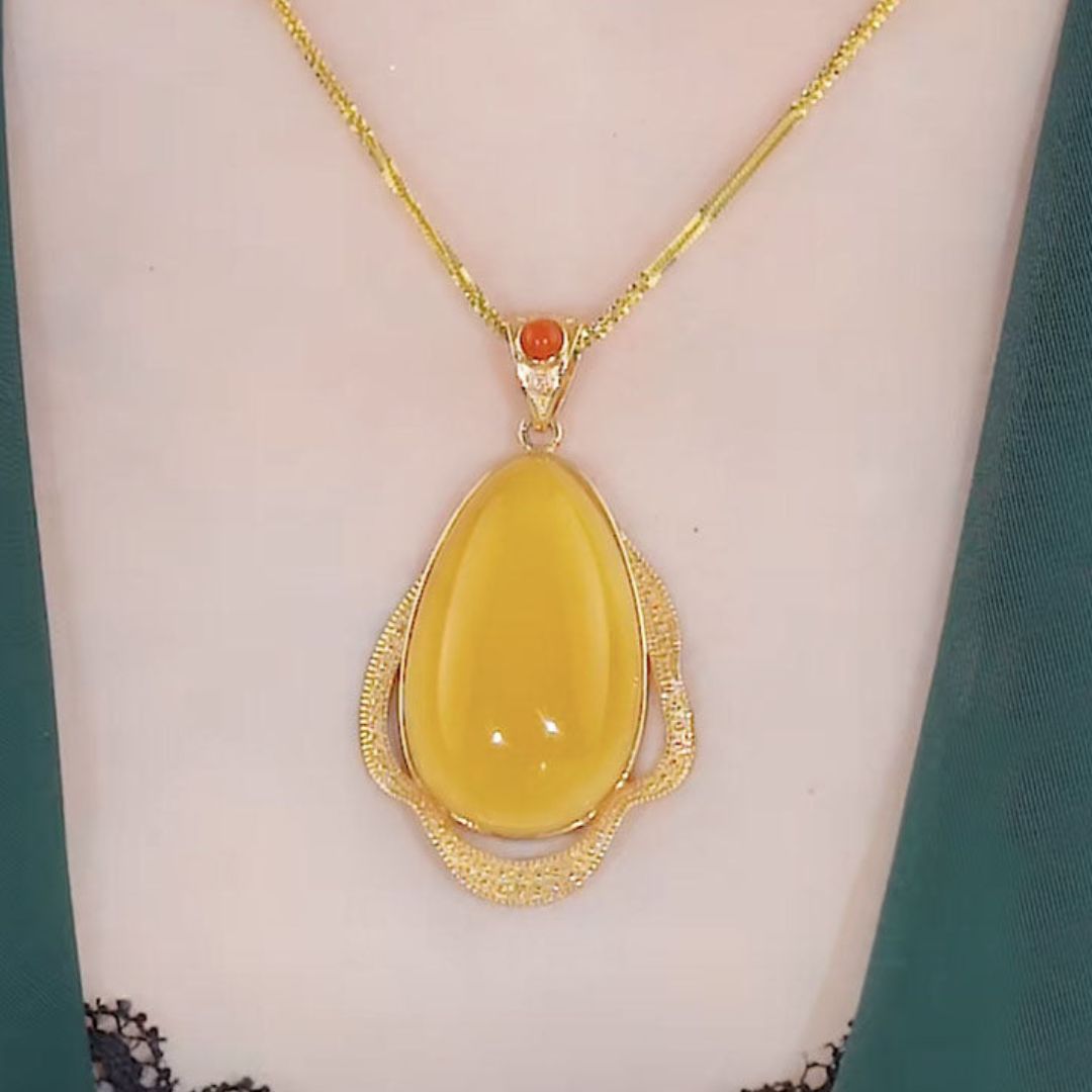 El collar amuleto de piedra ágata amarilla luce un gran dije ovalado enmarcado en oro texturizado y una pequeña piedra naranja, irradiando energía positiva sobre piel clara con prendas verde oscuro.