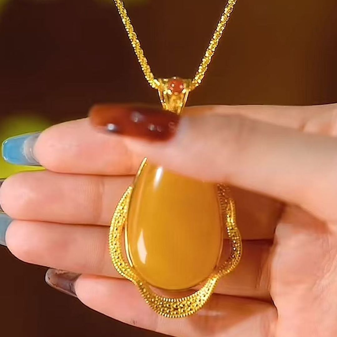 Una mano con uñas largas y decoradas sostiene un collar amuleto de piedra ágata amarilla: un collar de oro con una gran piedra de ágata amarilla en forma de lágrima enmarcada por un borde de oro texturado.