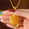 Una mano con uñas largas y decoradas sostiene un collar amuleto de piedra ágata amarilla: un collar de oro con una gran piedra de ágata amarilla en forma de lágrima enmarcada por un borde de oro texturado.