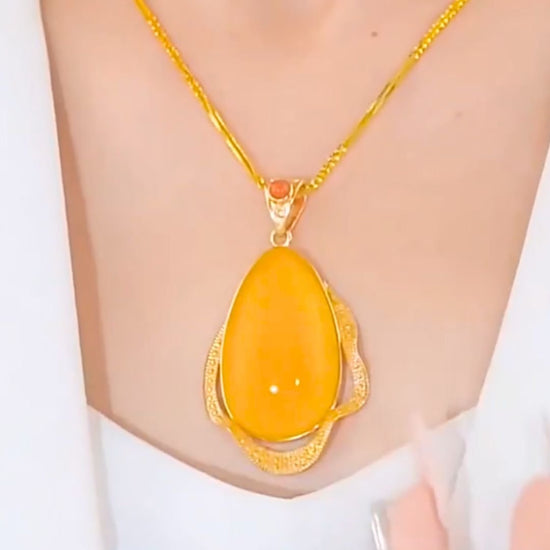 Primer plano de una persona que lleva el Collar amuleto de piedra ágata amarilla: un collar de oro con un gran colgante ovalado de piedra ágata amarilla con detalles decorativos dorados, sobre una prenda blanca.