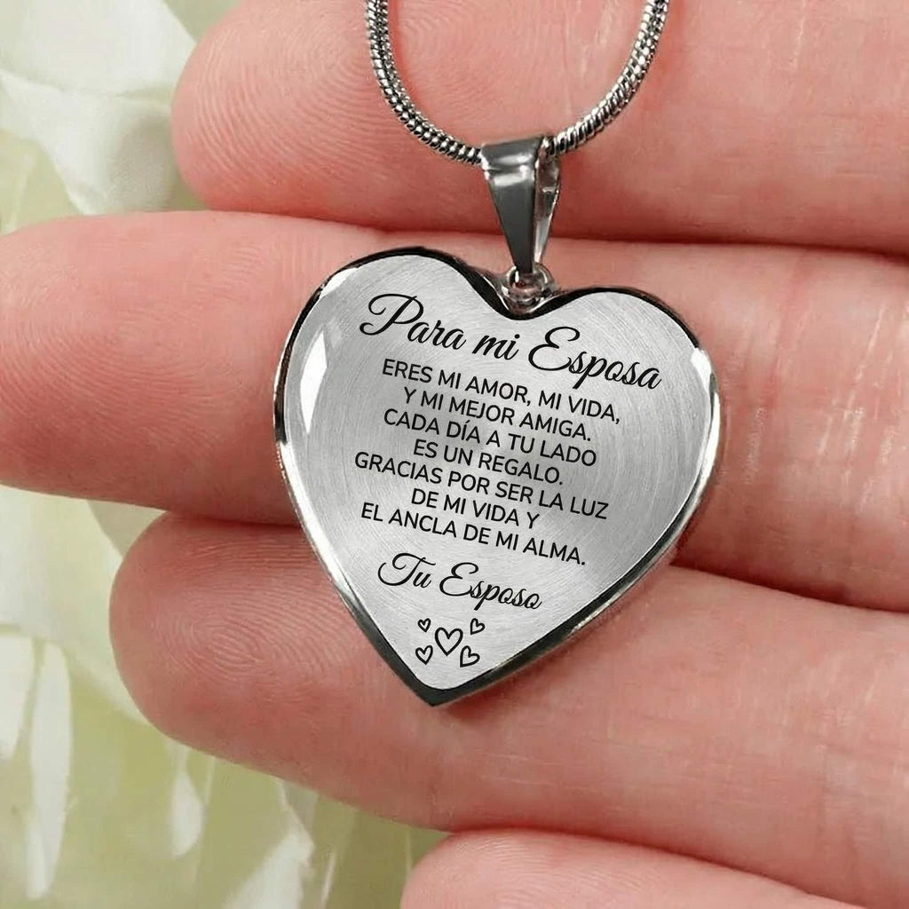 Una mano sostiene el "Collar con colgante Para mi esposa", un colgante de plata en forma de corazón grabado en español, que expresa amor y gratitud a una esposa, firmado "Tu Esposo" y decorado con pequeños corazones grabados.