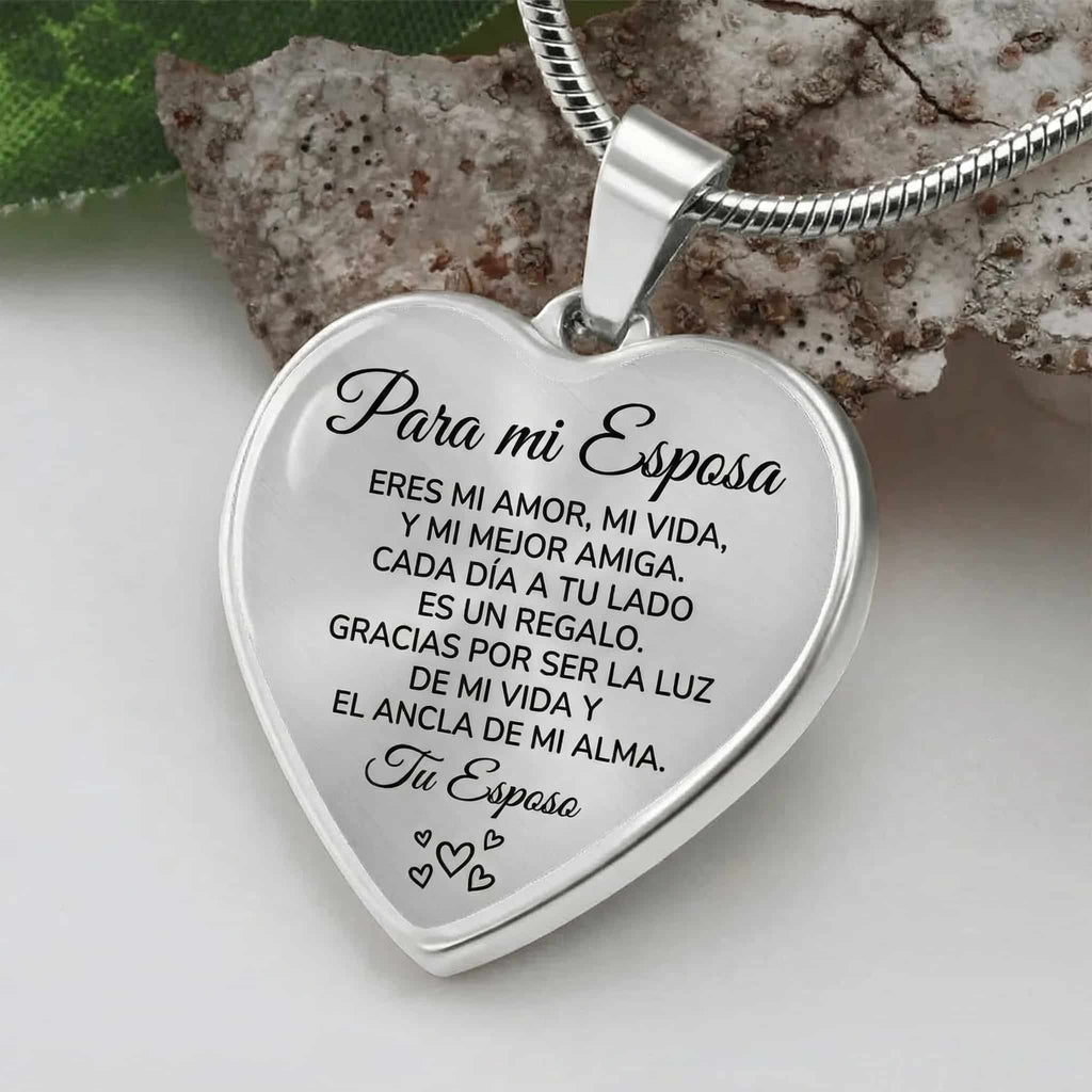 El collar con colgante "Para mi esposa", en forma de corazón plateado y grabado en español con un mensaje conmovedor, reposa sobre una piedra junto a hojas verdes, ideal como regalo especial de amor.