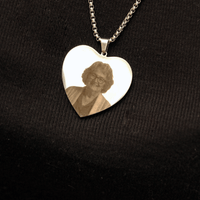 El collar con foto grabada en forma de corazón es un relicario de plata en forma de corazón con el retrato grabado de una anciana, que se muestra sobre una tela negra de canalé.