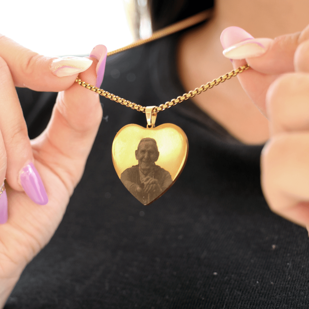 Una persona con uñas de color lavanda y blanco sostiene un Collar con foto grabada en forma de corazón, de oro, con la imagen grabada de una persona mayor. El fondo suavemente difuminado resalta esta pieza de joyería sentimental.