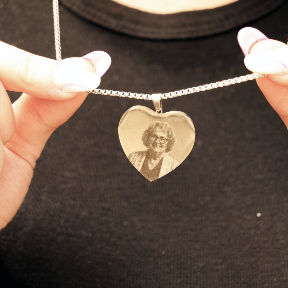 Alguien sostiene un Collar con foto grabada en forma de corazón -un colgante de plata en forma de corazón con un retrato grabado de una mujer mayor- sobre el fondo de una camisa negra.