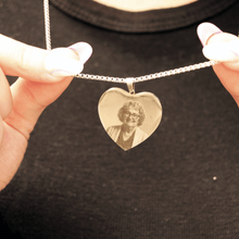 Cargar imagen en el visor de la galería, Alguien sostiene un Collar con foto grabada en forma de corazón -un colgante de plata en forma de corazón con un retrato grabado de una mujer mayor- sobre el fondo de una camisa negra.