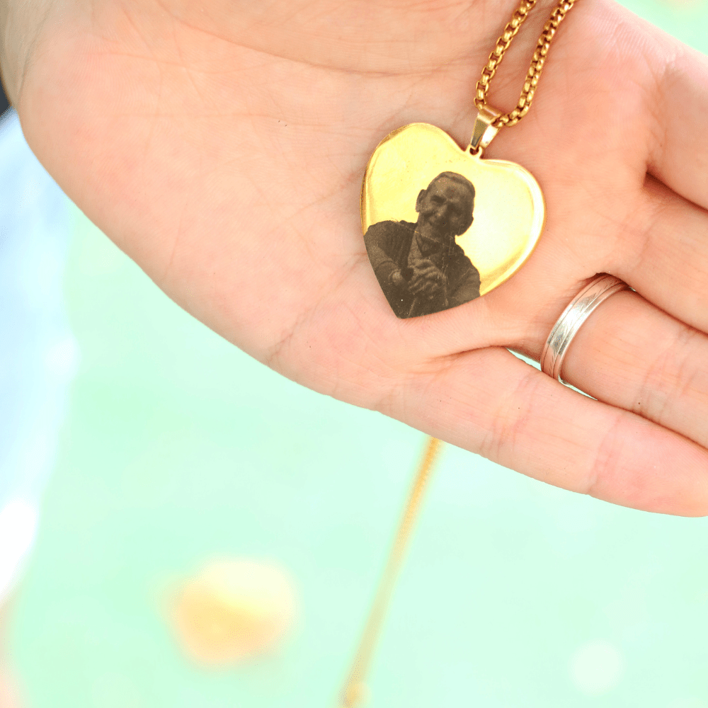 Una mano sostiene el Collar con foto grabada en forma de corazón, mostrando el retrato de un hombre. Quien lo sostiene lleva un anillo plateado y al fondo se aprecia un desenfoque verde claro.