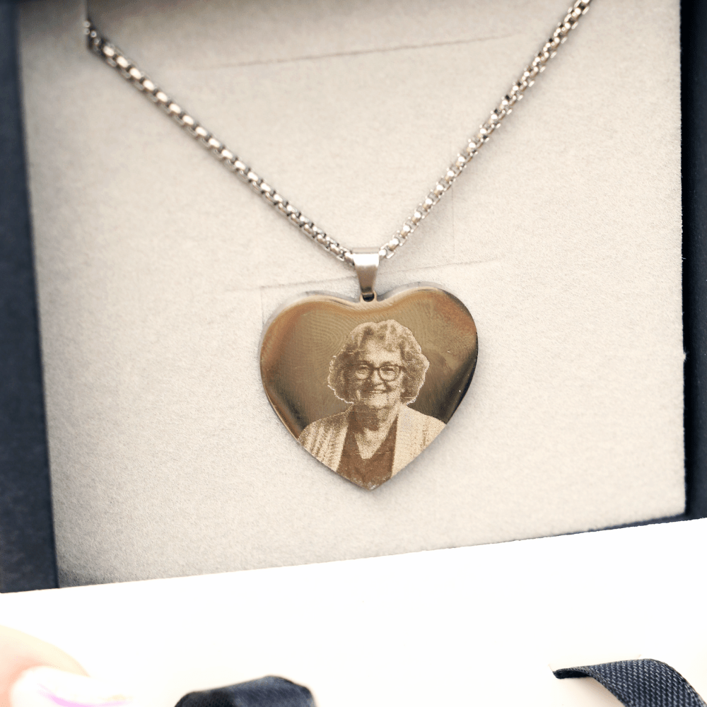 El Collar con foto grabada en forma de corazón es de plata e incluye el retrato grabado de una mujer mayor con gafas y cabello rizado, presentado en un estuche joyero con forro de tela clara.