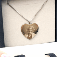 El Collar con foto grabada en forma de corazón es de plata e incluye el retrato grabado de una mujer mayor con gafas y cabello rizado, presentado en un estuche joyero con forro de tela clara.