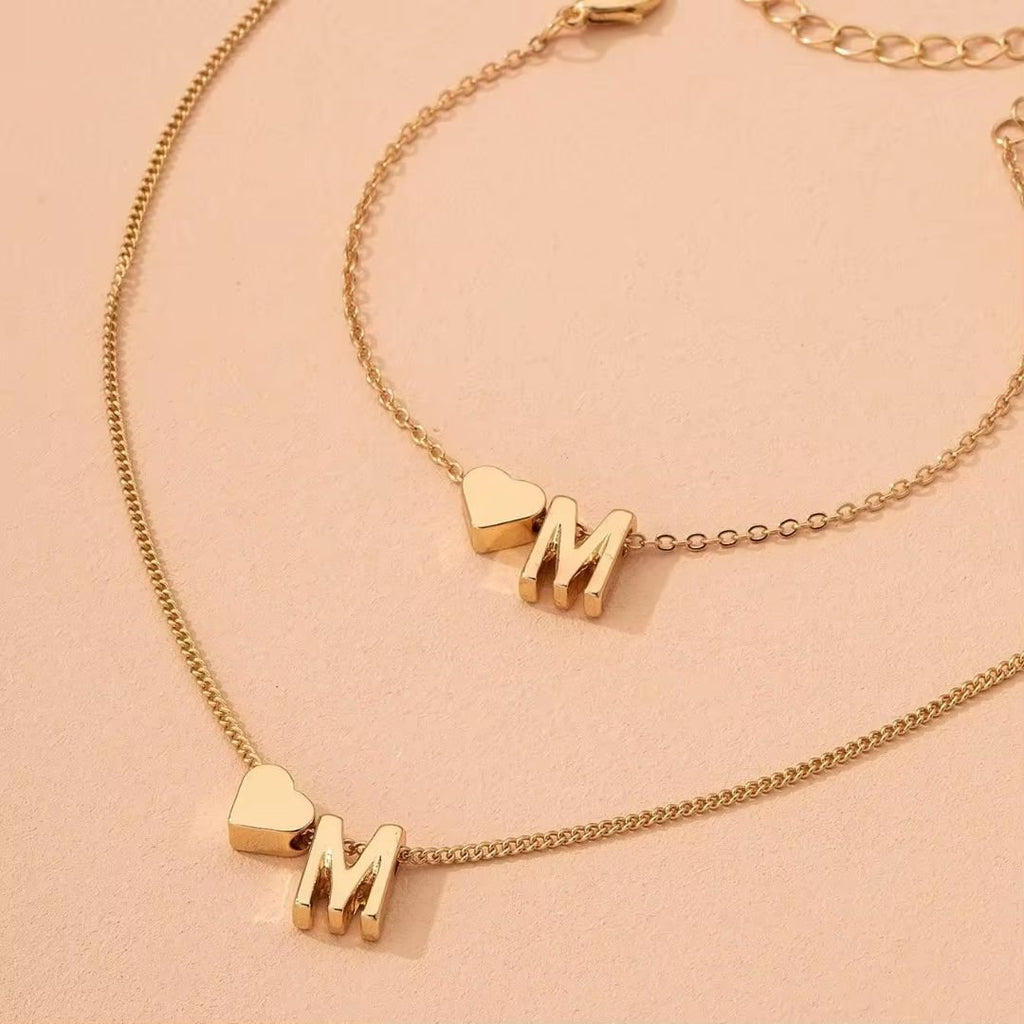 Dos collares dorados Collar con inicial y corazón: cada uno tiene un dije de corazón junto a la letra M, sobre una superficie beige. Perfectos como collar con inicial para un regalo personalizado.