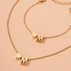Dos collares dorados Collar con inicial y corazón: cada uno tiene un dije de corazón junto a la letra M, sobre una superficie beige. Perfectos como collar con inicial para un regalo personalizado.