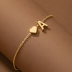 Un delicado collar dorado con inicial y corazón que incluye un pequeño colgante en forma de corazón y la letra A. Se lleva sobre un fondo marrón neutro y es perfecto como regalo personalizado.