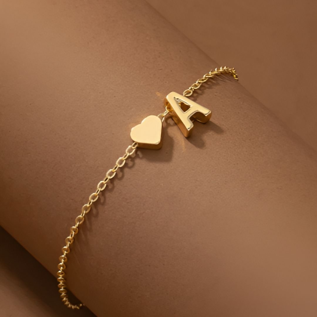 Un delicado collar dorado con inicial y corazón que incluye un pequeño colgante en forma de corazón y la letra A. Se lleva sobre un fondo marrón neutro y es perfecto como regalo personalizado.