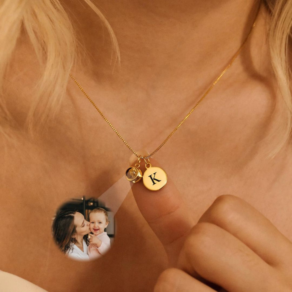 Una persona usa un Collar con inicial y foto en color dorado, con dije redondo grabado con la letra K y charm de piedra, sosteniendo el dije. En una imagen pequeña, una mujer besa a un bebé sonriente. Ideal para quienes aman los collares personalizados.
