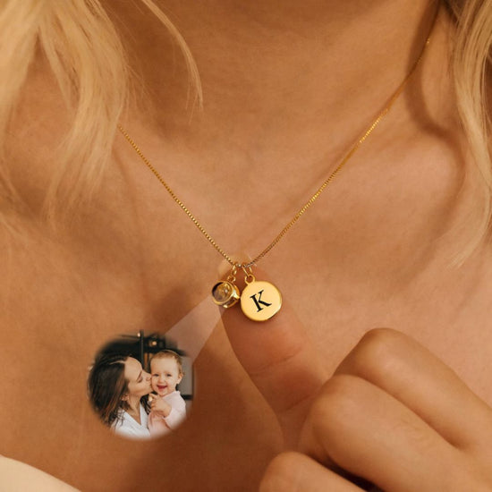 Una persona usa un Collar con inicial y foto en color dorado, con dije redondo grabado con la letra K y charm de piedra, sosteniendo el dije. En una imagen pequeña, una mujer besa a un bebé sonriente. Ideal para quienes aman los collares personalizados.