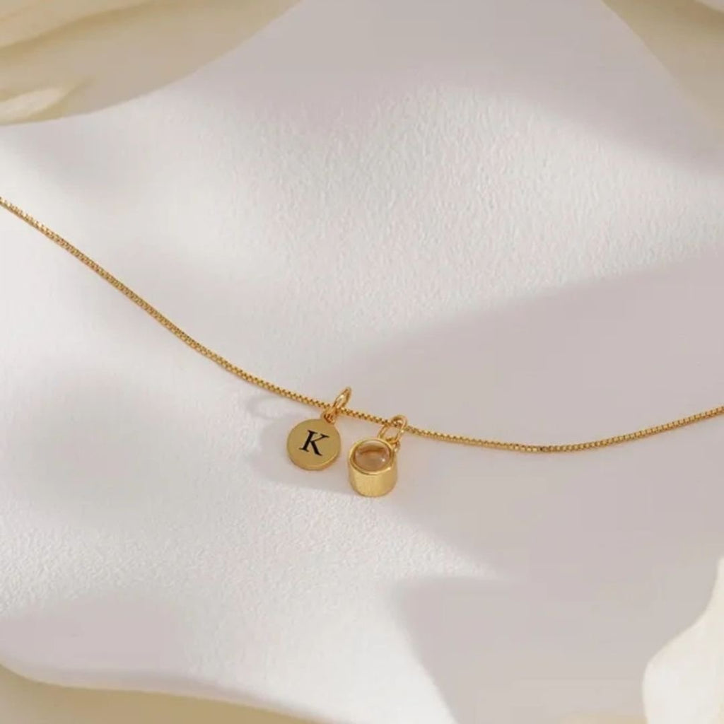 El "Collar con inicial y foto", un delicado collar de oro con un disco redondo grabado con la letra K y un pequeño anillo de oro, capta maravillosamente el encanto de la joyería personalizada, expuesta con elegancia sobre una suave superficie blanca.